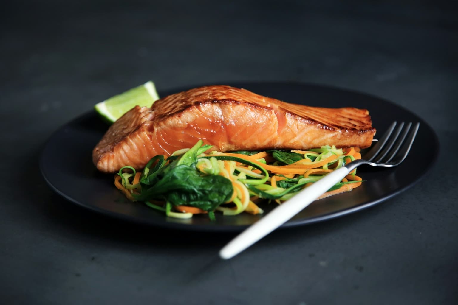 A seared fish fillet plated on ribboned vegetables with a wedge of lime